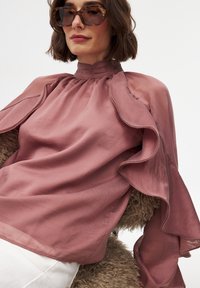 Roze blouse met gerimpelde mouwen, lichte stof, hoge halslijn en een gladde textuur. Gekocht over een gestructureerd oppervlak.