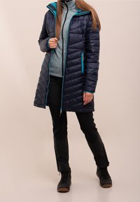 Marineblå pufferjakke med turkise detaljer, quiltet design, lynlåslommer og aftagelig hætte, båret over en grå fleece-trøje og sorte bukser.