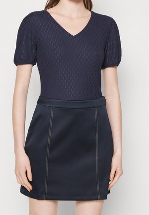 Kobieta ubrana w granatowy top z krótkim rękawem i dekoltem w serek o fakturowanej tkaninie, połączony z gładką granatową spódnicą w kształcie litery A z pionowymi przeszyciami.