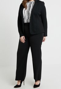Zwarte blazer met een aangesloten ontwerp, gecombineerd met zwart wijduitlopende broek. De top heeft verticale zwart-witte strepen. Zwarte hakken maken de look compleet.