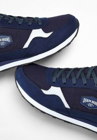 Sneakers blu navy con tomaia in suede e tessuto, dettagli bianchi, lacci piatti e un'etichetta con il marchio sul lato. Suola in gomma per una maggiore aderenza.