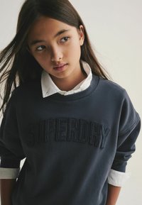 Navyblaues Sweatshirt mit dem aufgestickten Schriftzug "SUPERDRY" in Dunkelblau, kombiniert mit einem weißen Hemd, das aus dem Bund hervorschaut.