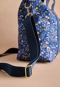 Blå tygväska med färgglada små blommönster och husmotiv, utrustad med en marinblå justerbar rem samt spänne och lås i guldfärgad metall.
