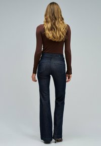 Jeans acampanados de mezclilla azul oscuro con cintura alta, dos bolsillos traseros y una textura suave, combinado con una blusa ajustada de mangas largas color marrón.