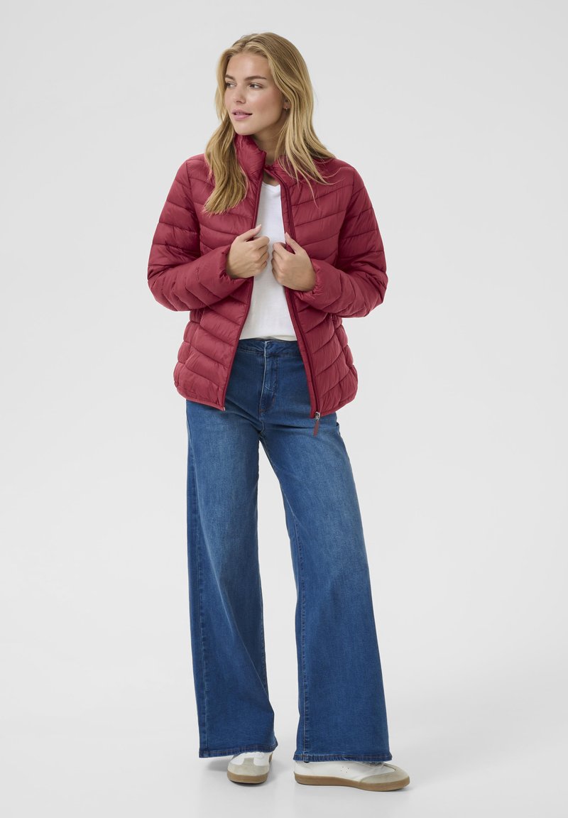 Giacca puffer borgogna con texture trapuntata e colletto alto, abbinata a jeans a gamba larga blu chiaro e sneakers bianche su uno sfondo neutro.