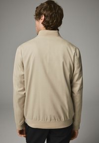 Beige jakke med ståkrave, struktureret stof og ribbede manchetter. Har en glat overflade og elastisk talje i kanten.