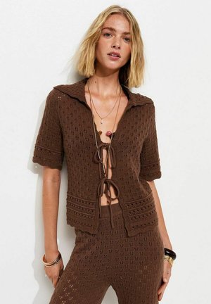 Femme blonde portant un haut court en tricot marron à manches courtes avec des fermetures à nouer et un pantalon taille haute assorti, accessoirisée de bracelets et de colliers.