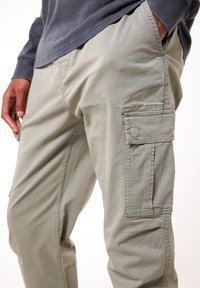 Vaaleanharmaat cargo-housut, jotka on valmistettu pehmeästä kankaasta. Housuissa on sivutaskut ja näkyvät ompeluyksityiskohdat, rento leikkaus ja hieman kiiltävä pinta.