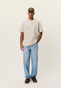 Μπεζ υπερμεγέθη μπλουζάκι, ανοιχτό μπλε τζιν με ίσια γραμμή και καφέ παπούτσια. Καπέλο ελιάς με λευκά γράμματα στο μπροστινό μέρος.