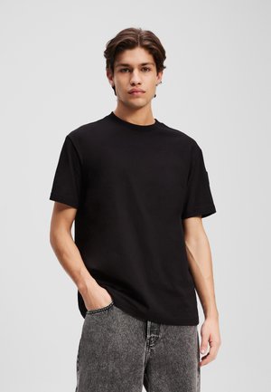 T-shirt nera in cotone con maniche corte e collo a giro, caratterizzata da un fit rilassato e un design minimale, abbinata a jeans grigio scuro.