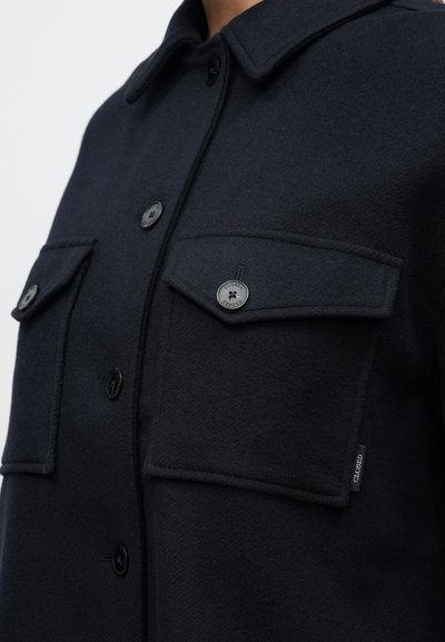 Ναυτικό μάλλινο πουκάμισο-ζακέτα με μυτερό γιακά, δύο τσέπες στο στήθος, μαύρα κουμπιά και ετικέτα στο πλάι. Λείο ύφασμα με καθαρές γραμμές.