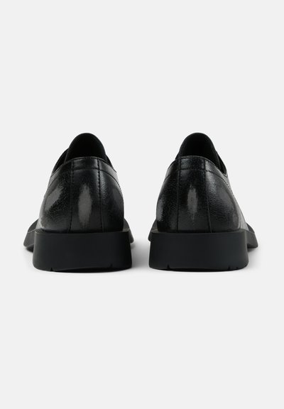 Ett par svarta läderloafers sedda bakifrån med låg klack och en slät, svagt glänsande yta på en enkel bakgrund.