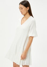 Robe blanche avec un décolleté en V, des manches courtes bouffantes et un ourlet à volants superposés. Un tissu texturé avec un motif subtil ajoute de la dimension.