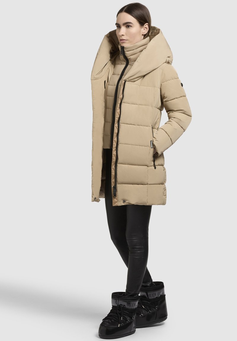 Beiger Puffer-Mantel mit großem Kragen, Reißverschluss und seitlichen Taschen. Kombiniert mit schwarzen Leggings und schwarzen Schnürstiefeln. Glatte Textur.