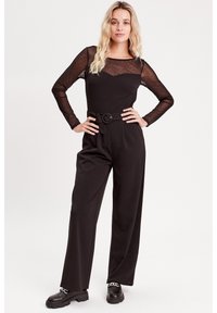Combinaison noire avec des manches longues en tulle à pois, un corsage ajusté, un pantalon à jambes larges et une taille ceinturée. Des chaussures noires épaisses complètent le look.