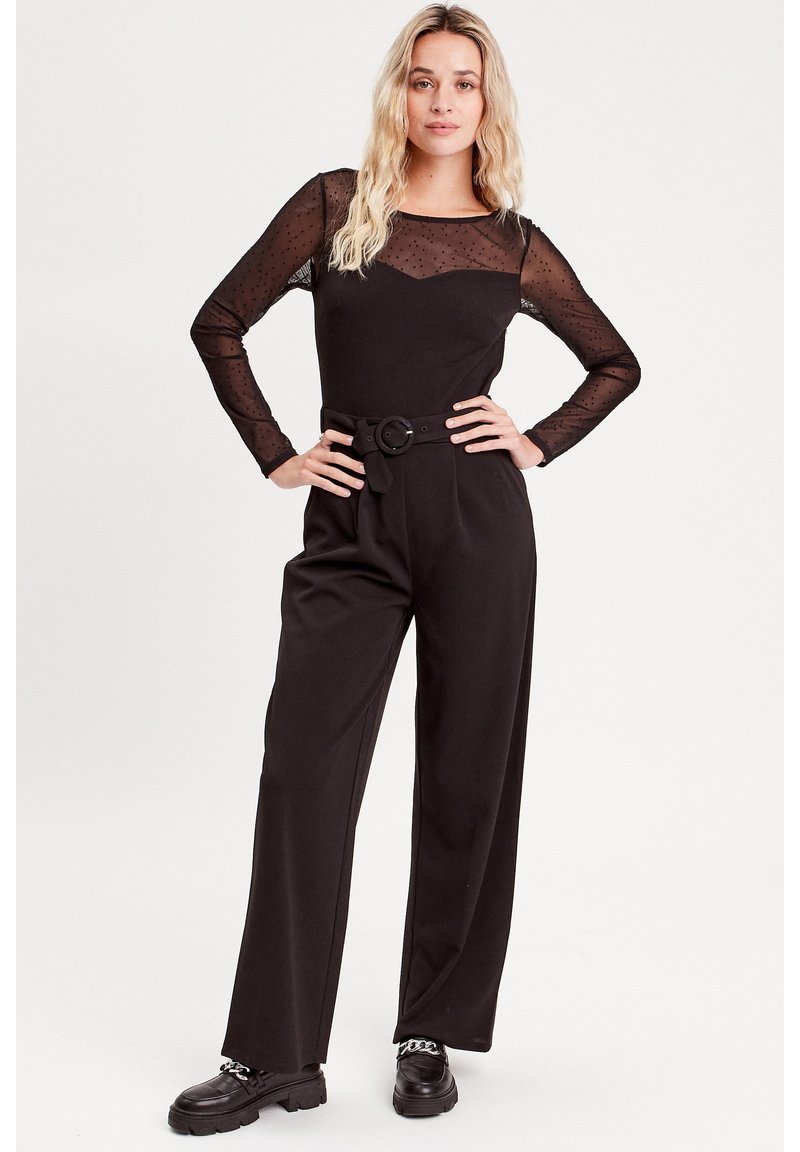 Combinaison noire avec des manches longues en tulle à pois, un corsage ajusté, un pantalon à jambes larges et une taille ceinturée. Des chaussures noires épaisses complètent le look.