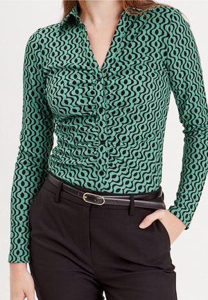 Mujer vistiendo una camisa verde de manga larga con botones y un patrón de puntos ondulados negros, pantalones negros y un cinturón negro, con la mano en el bolsillo.