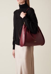 Maroon leather handbag with a textured edge, softly rounded shape, and a gold logo. It is held by a person wearing a black sweater.