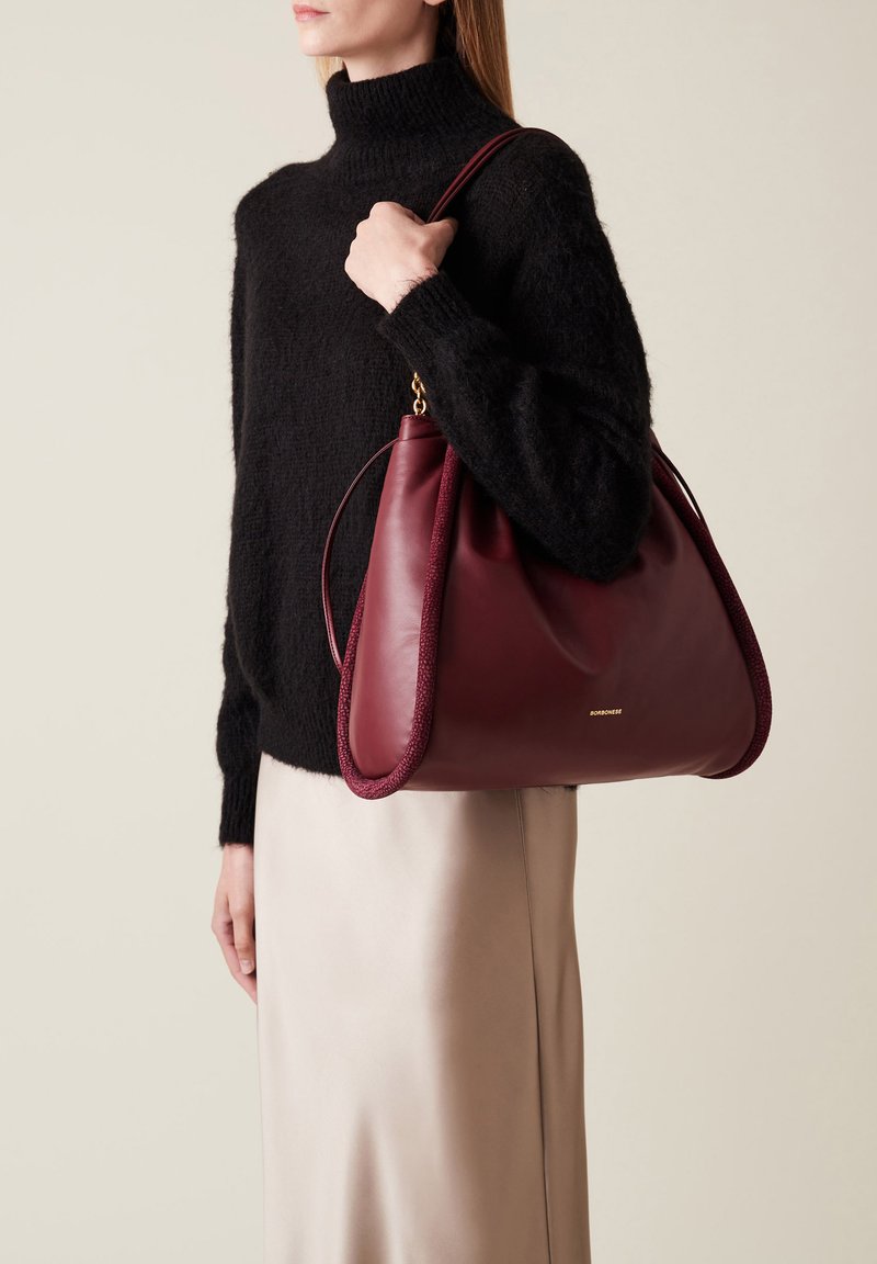 Maroon leather handbag with a textured edge, softly rounded shape, and a gold logo. It is held by a person wearing a black sweater.