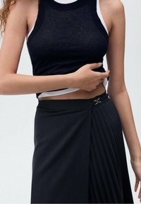 Débardeur noir côtelé superposé sur un haut blanc, associé à une jupe plissée noire avec un accent en matériel doré à la taille.