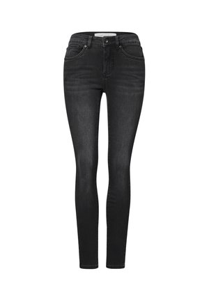 Kvinde iført sorte jeans og en leopardprint top, der holder en bog med den ene hånd, mens den anden hånd hviler på hendes hofte.