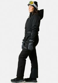 Zwart skikostuum met een aangesloten ontwerp, verstelbare riem en handschoenen. Voorzien van een capuchon en reflecterende elementen. Draagt ski-goggles en zwarte laarzen.