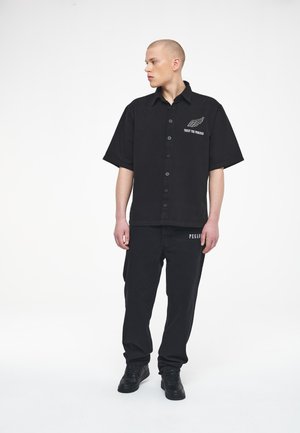 Young man with buzz cut wearing black button-up shirt with "Trust The Process" text and black pants with "Pegador" text, standing against white background.