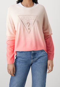 Pull léger en dégradé de rose, avec un logo triangulaire central et un texte en relief. Manches longues, coupe décontractée et texture douce.