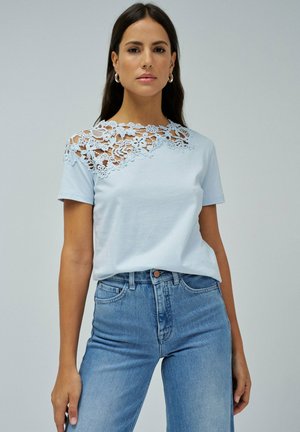 Camiseta azul claro con detalles de encaje floral en el hombro izquierdo y mangas cortas, combinada con jeans de mezclilla azul claro.