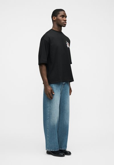 Άνδρας όρθιος, φοράει μαύρο υπερμεγέθες μπλουζάκι με σχέδιο, φαρδιά ανοιχτό μπλε τζιν και μαύρα παπούτσια, μπροστά σε απλό λευκό φόντο.