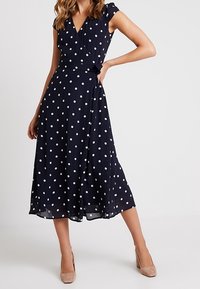 Granatowa sukienka kopertowa w białe kropki, z krótkimi rękawami, paskiem w talii, lejącą się spódnicą sięgającą do połowy łydki. W zestawieniu z beżowymi szpilkami.