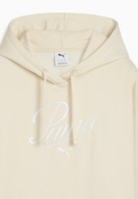 Béžová mikina Puma s kapucňou na šnúrku, so bielym vyšívaným logom a textom na prednej strane. Hladká štruktúra a voľný strih.