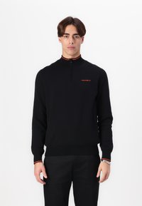 Suéter de punto negro con cuello alto, cierre de cremallera en la parte frontal y acentos rojos. Presenta el logo "HACKETT" en el pecho, puños ajustados y dobladillo.