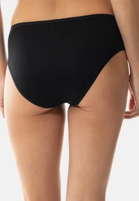 Eine Person trägt schlichte schwarze hochgeschnittene Unterwäsche, gezeigt von der unteren Rückseite bis zur Mitte des Oberschenkels vor einem schlichten weißen Hintergrund.