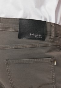 Pilkos medžiagos kelnės su juodu odiniu pledu, ant kurio išreikštas „Jack & Jones“ logotipas. Yra standartinė užpakalinė kišenė ir siūlių detalės.