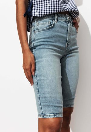 Personne portant un short en jean bleu clair jusqu'aux genoux et une chemise à carreaux bleue et blanche, debout avec une main reposant sur le côté.