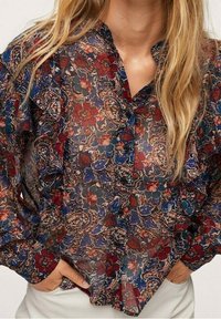 Blouse transparente à manches longues avec des motifs floraux et paisley rouges, bleus et marron, portée à l'intérieur d'un pantalon blanc avec les mains dans les poches.
