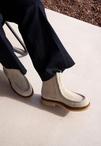 Béžové kotníkové boty z béžové semiše s elastickými bočními panely, zaoblenou špičkou a strukturovaným povrchem. Hnědé gumové podrážky poskytují kontrast a odolnost.