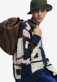 Häkelpullover mit dunkelblauen, grünen, rosa und cremefarbenen Quadraten; gerippte Bündchen; kombiniert mit einer braunen Ledertasche mit Streifen und einem Griff.