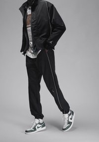 Svart zip-up jacka med vit piping, svarta byxor och vita/gröna sneakers. T-shirten har en grafisk design i jordnära färger.