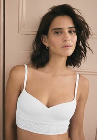 Bralette bianco con design a scollo a V, spalline sottili e una fascia in pizzo sul fondo. Materiale liscio con una texture morbida.