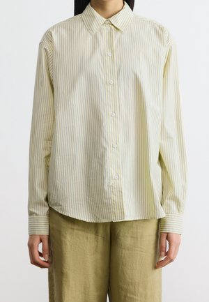 Personne portant une chemise jaune clair à manches longues boutonnée avec de fines rayures verticales et un pantalon beige ample, debout devant un fond uni.