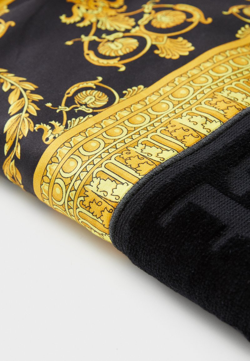 Schwarzes Handtuch mit kunstvollen goldenen Blumenmustern und Bordüren, weichem Teppichgefühl und einem markanten H-Muster in Schwarz am Rand.