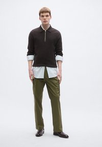 Brūns džemperis ar rāvējslēdzēja kaklu pār baltu svītrainu kreklu un olīvzaļas krāsas kargo bikses. Brūnas pulētas kurpes, brīvs piegriezums, mīkstas faktūras, minimālistisks dizains.