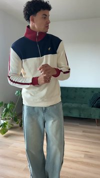 Felpa blu navy e crema con zip, dettagli bordeaux e polsini a coste, abbinata a jeans a gamba larga azzurri. Ambientazione interna con divano verde.