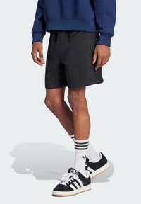 Schwarze Baumwollshorts mit seitlichen Taschen und dezentem Logo, kombiniert mit schwarzen Sneakers mit weißen Streifen und weißen Socken mit schwarzen Akzenten.