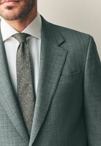 Šedý texturovaný oblekový sako s klopou, bílé košile a světlé šedé tkané kravaty. Viditelná kapsa na levé straně saka.