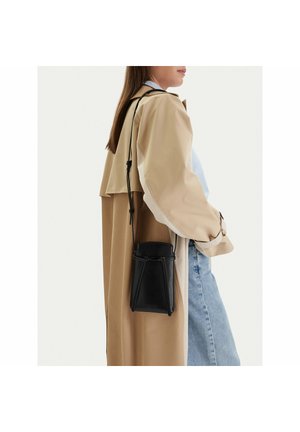 Žena v béžovém trenčkotě a světle modrých džínách nosí na rameni malou černou koženou crossbody kabelku s tenkým páskem.