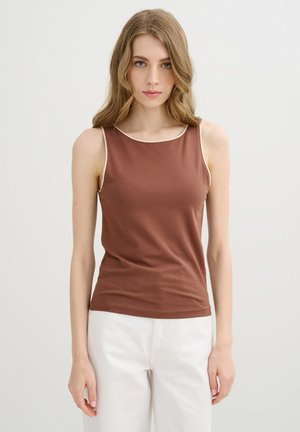 Joven mujer con cabello rubio ondulado que lleva una camiseta marrón sin mangas y pantalones blancos, de pie frente a un fondo claro y liso.