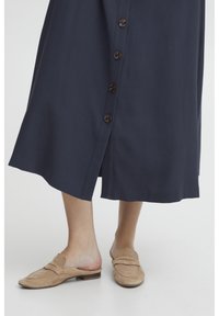 Abito blu navy a lunghezza ginocchio con chiusura a bottoni, caratterizzato da bottoni marroni e una silhouette fluida. Abbinato a mocassini in suede color talpa.